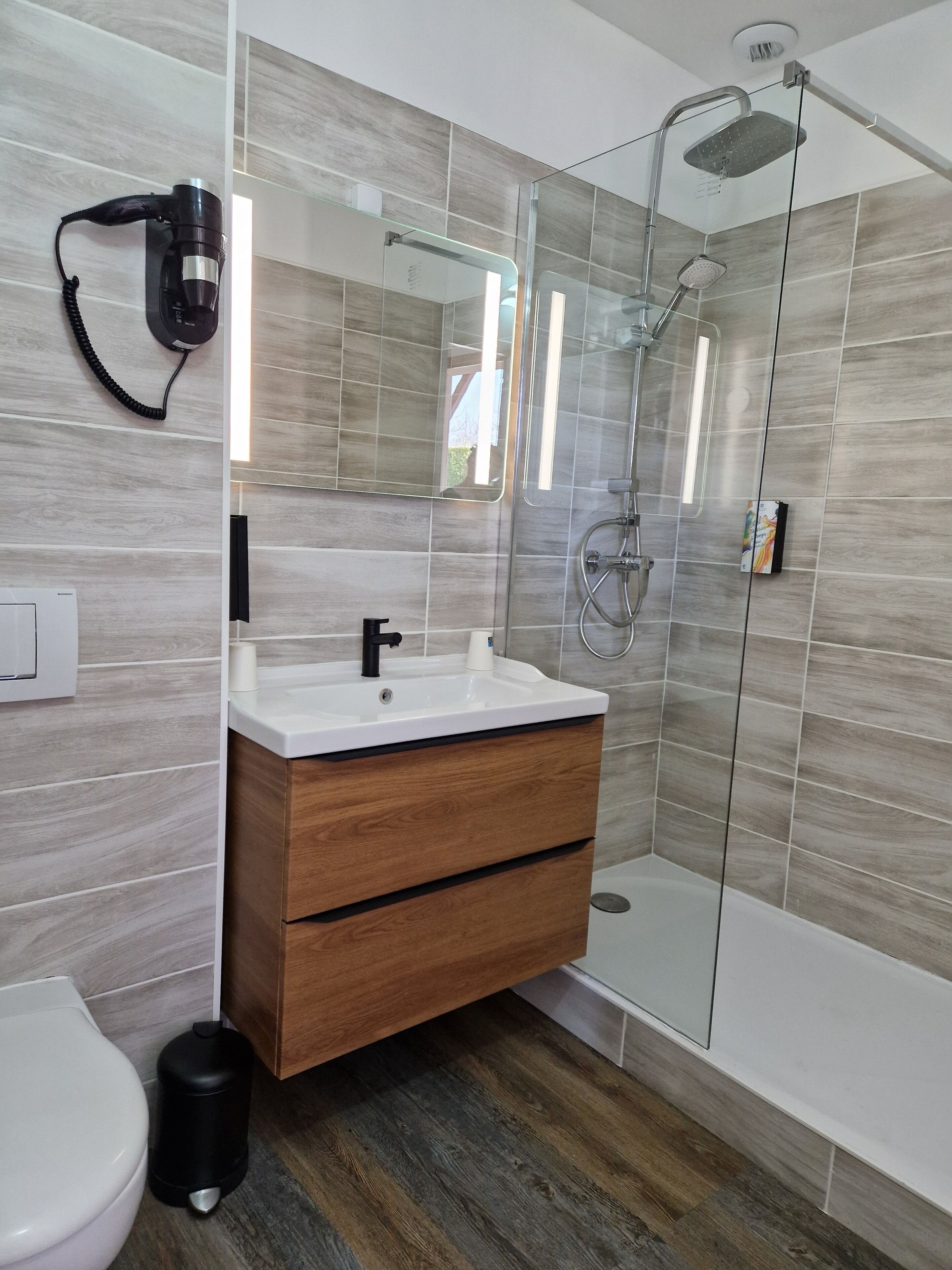 Une salle de bain moderne avec un meuble-lavabo en bois, une cabine de douche en verre et un sèche-cheveux fixé au mur.