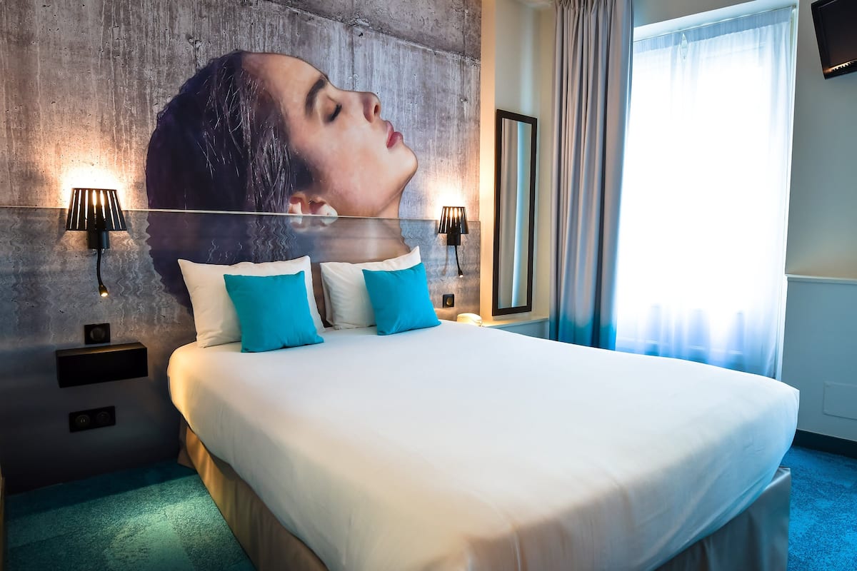 Une chambre d’hôtel avec un grand lit, deux lampes fixées au mur, une fenêtre avec des rideaux et un mur à motifs.