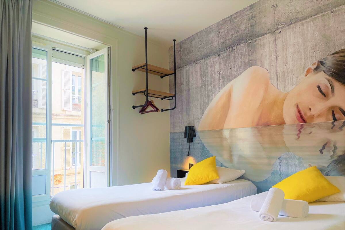 Une chambre d’hôtel avec deux lits, une grande fenêtre, un porte-serviettes fixé au mur et un tableau.