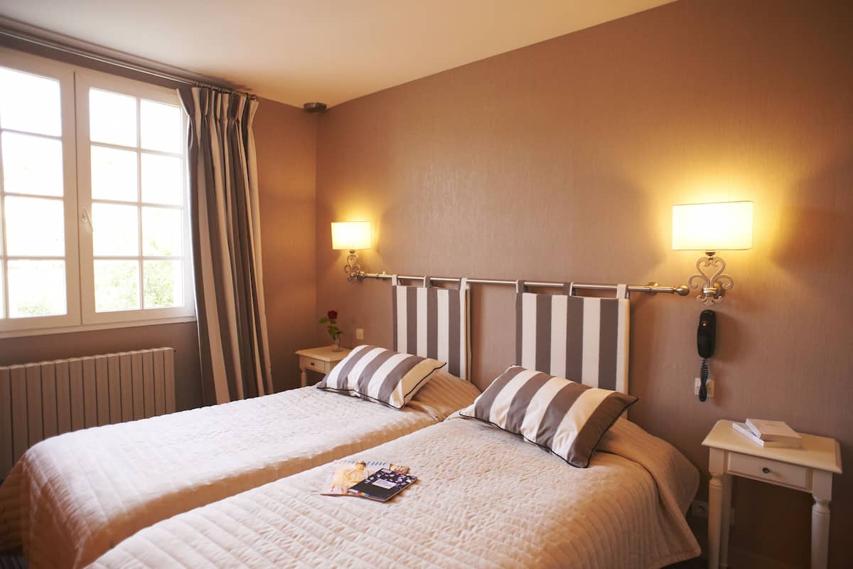 Une chambre d’hôtel avec deux lits, une fenêtre avec des rideaux, des lampes de chevet, une table de chevet et un téléphone fixé au mur.