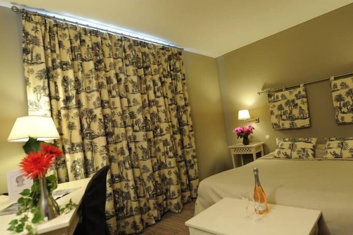 Une chambre d’hôtel avec un lit, un bureau, une chaise, une lampe, un vase avec des fleurs et une bouteille sur une petite table.