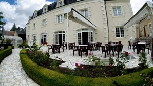 A large building with multiple windows and doors, outdoor seating areas with tables and chairs, and a well-maintained garden with flowers and hedges.