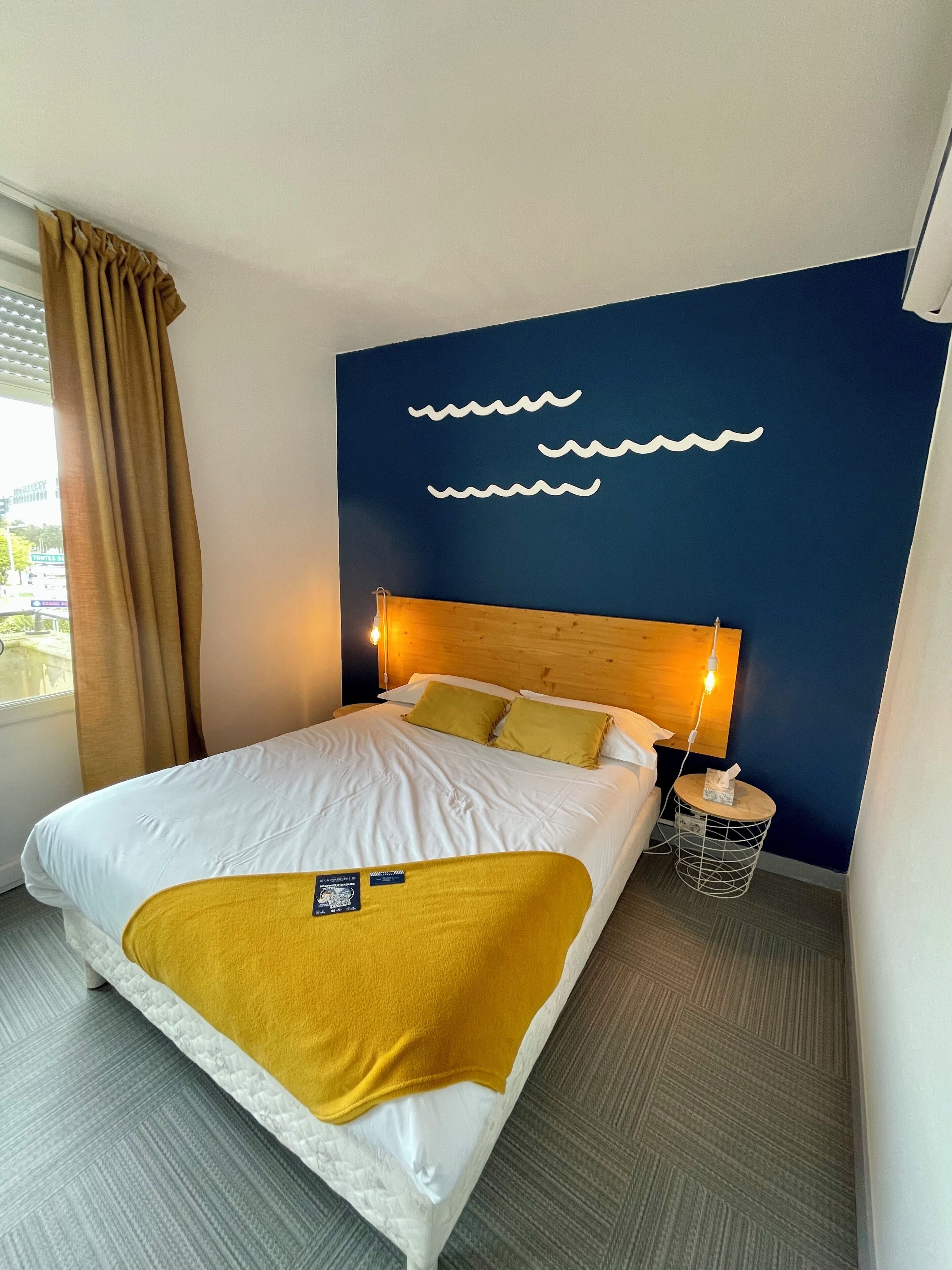 A hotel room with a bed, a headboard, a nightstand, a wall with a wave design, and a window with curtains.