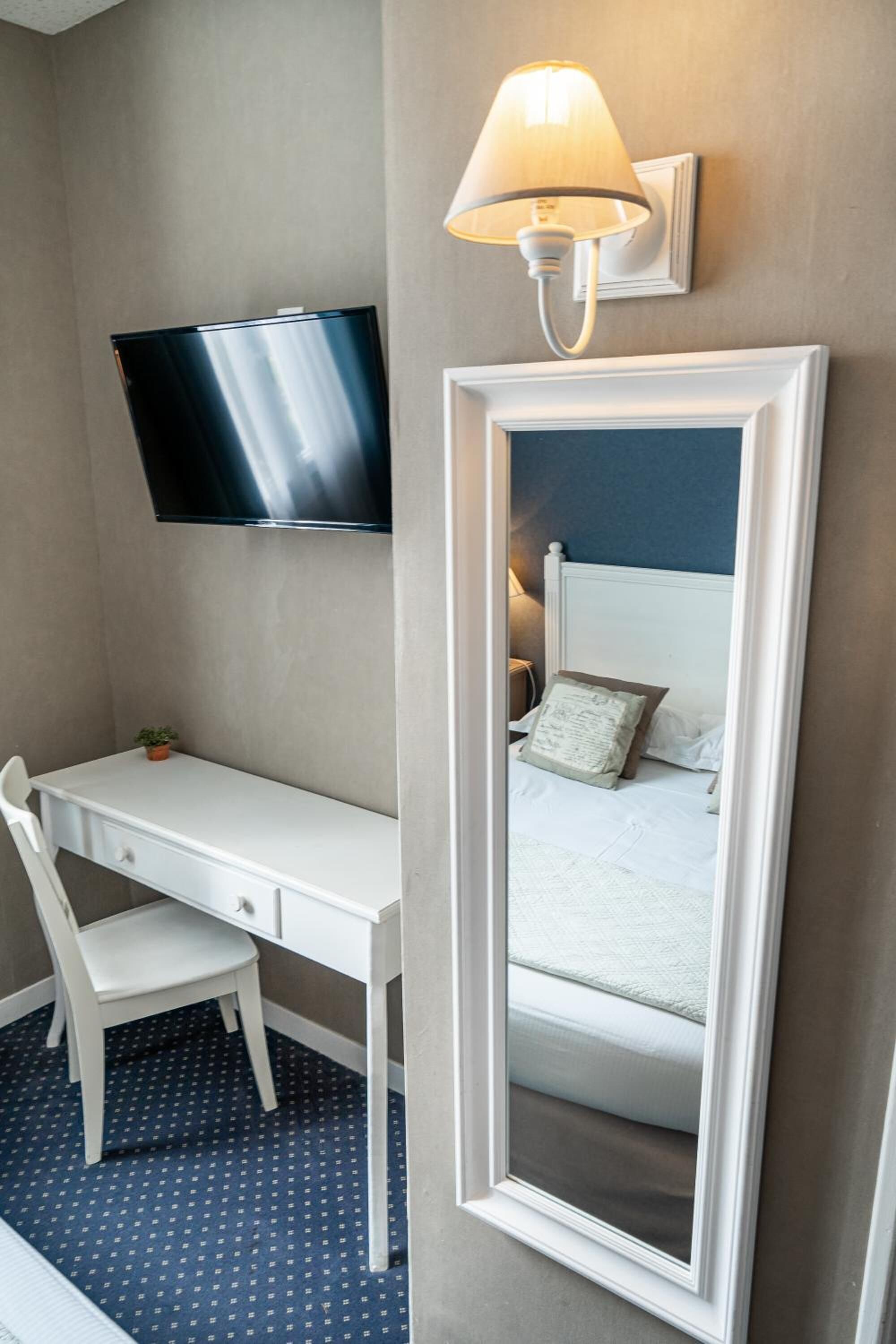A hotel room with a bed, a desk with a chair, a television, and a wall-mounted lamp.