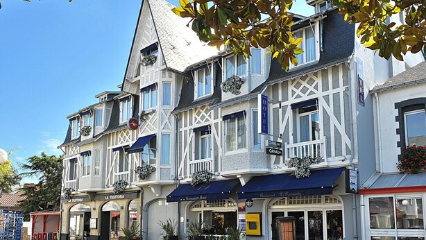 Un hotel europeo tradicional de varios pisos, con balcones adornados con macetas de flores y un letrero que indica un restaurante llamado "Le Verandah" en la planta baja.