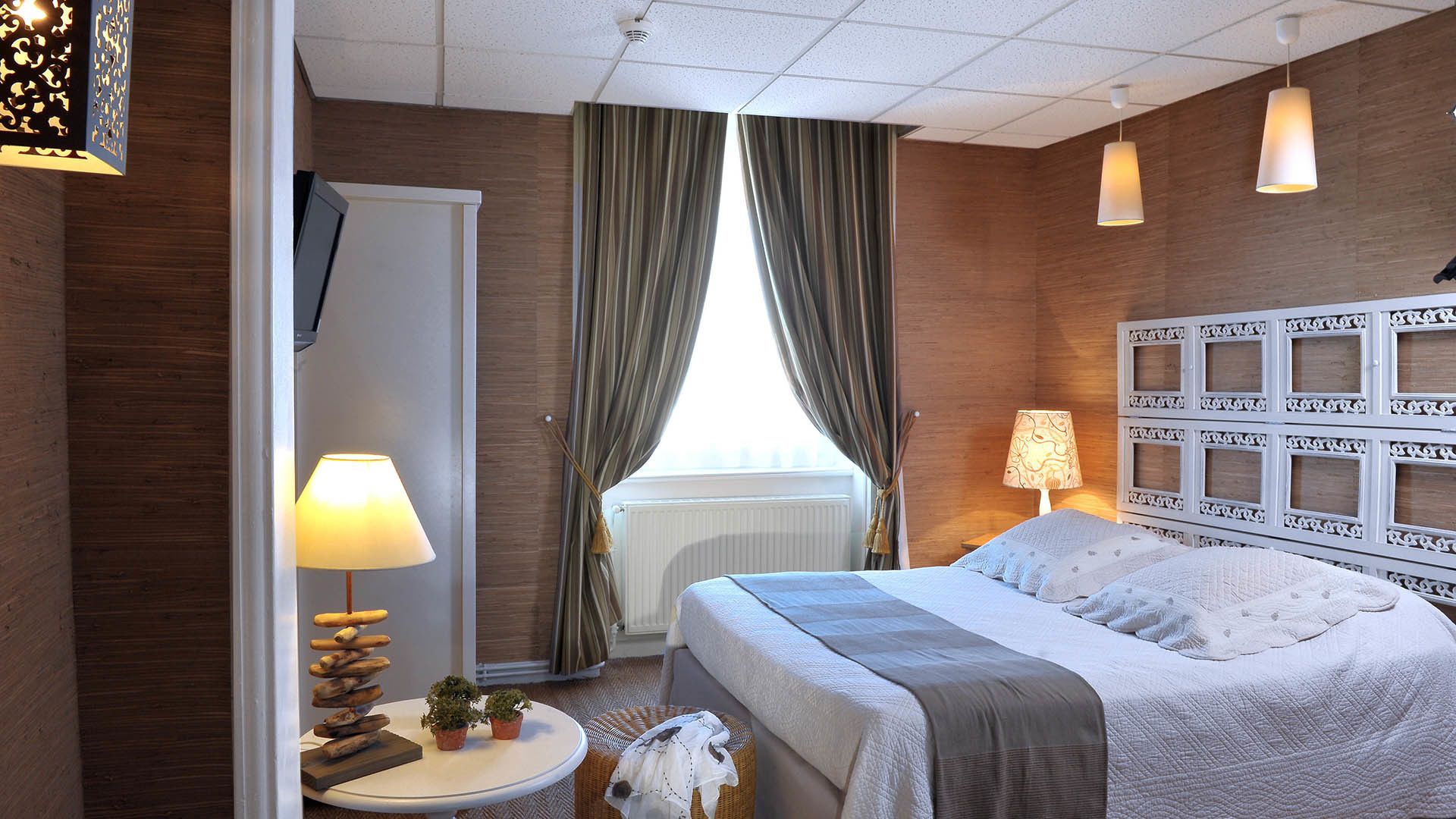 Habitación de hotel con cama, dos lámparas de noche, un cabecero decorativo, una mesita redonda con lámpara, una planta pequeña y un televisor montado en la pared.