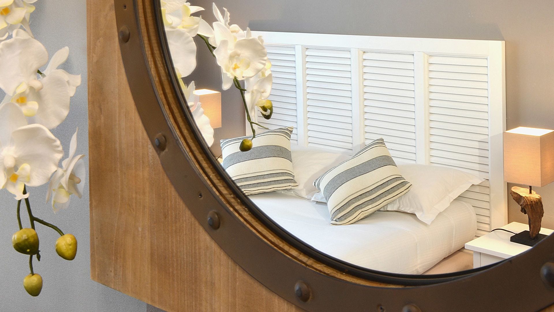 Ein Schlafzimmer mit Bett, Kissen, Nachttisch, Lampe und einem Spiegel mit Orchideen.
