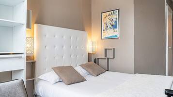 Una habitación de hotel con un cabecero capitonado, una cama con ropa de cama blanca y marrón, un cartel colgado en la pared y una mesita de noche con una lámpara.