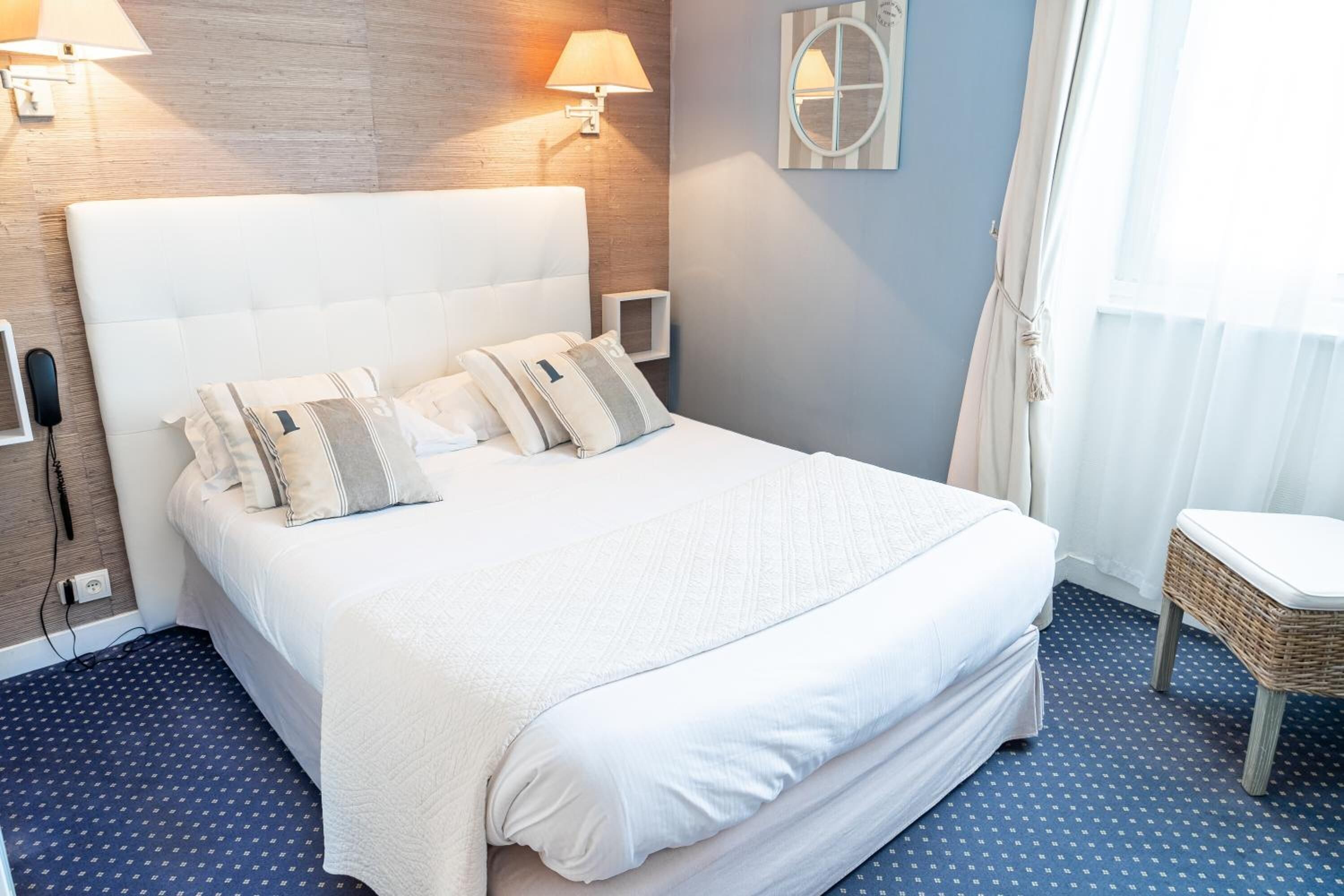 Una cama bien hecha con cabecera blanca y varios almohadones, una mesita de noche con una lámpara y una silla de mimbre.