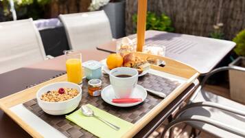A breakfast tray with a bowl of cereal, a glass of orange juice, a cup of coffee, and a croissant on a wooden table outdoors.