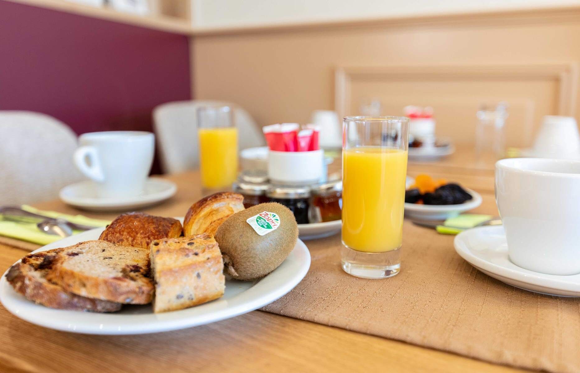 A breakfast setting with toast, kiwi, orange juice, and coffee cups on a table.