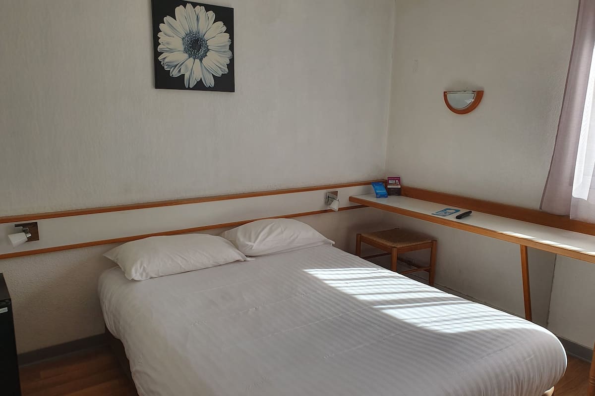 Una habitación de hotel con una cama, un escritorio, una silla, un espejo adosado a la pared y un cuadro de una sola flor.