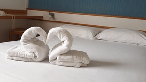 Una cama bien hecha con dos toallas dispuestas en forma de cisne, una mesita de noche de madera con un teléfono, y un cuadro en la pared.