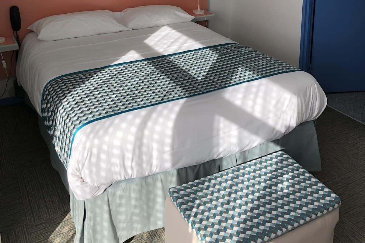 A hotel room with a bed, a bedside table with a lamp, a wall-mounted painting, and a blue door.