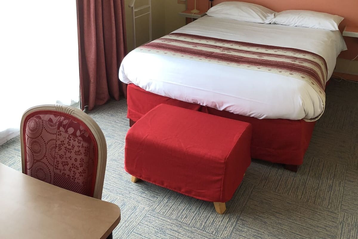 A hotel room with a bed, a bedside table, a chair, a painting on the wall, and a window with curtains.