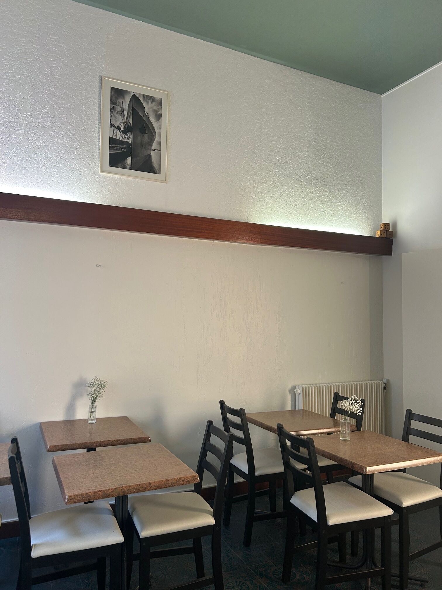 Een eethoek met houten tafels en stoelen, een lijst met een foto aan de muur, en een radiator eronder.