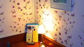 A corner room with a wooden table, two blue chairs, a lit table lamp, a framed painting on the wall, and a floral wallpaper design.