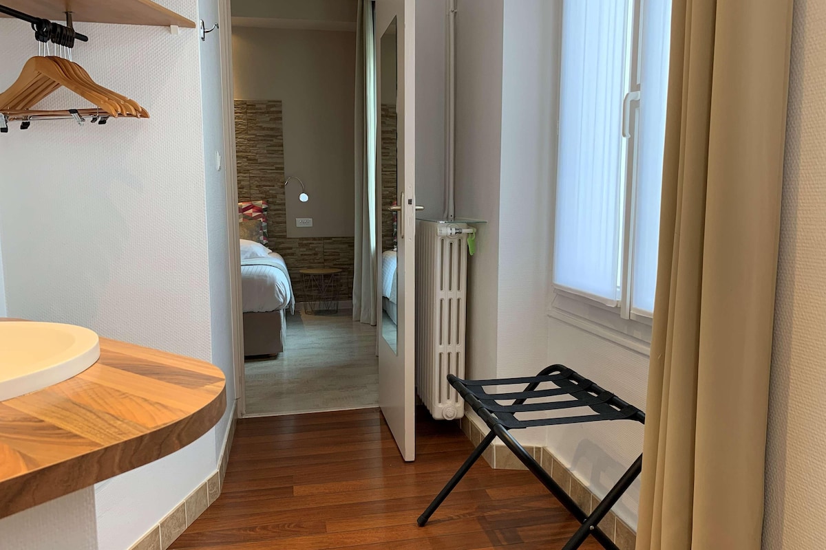 Ein Hotelzimmer mit einem Holznähtisch, einem Heizkörper, einem Bett mit geometrischem Kopfstück und einem Fenster mit Vorhängen.
