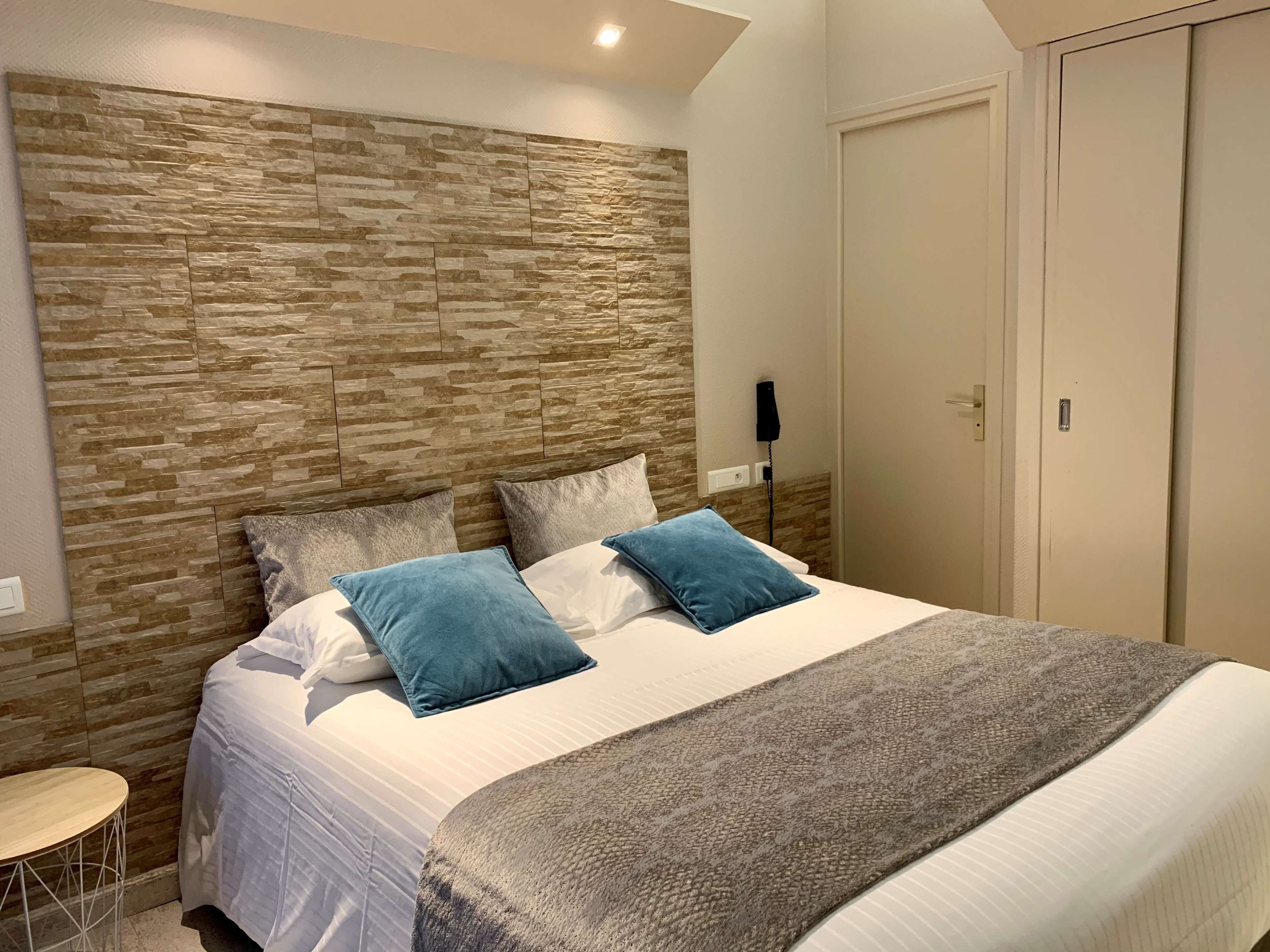 Un dormitorio con cabecera estampada en piedra, cama con ropa de cama blanca y dos almohadas azules, una manta gris y una pequeña mesa redonda.