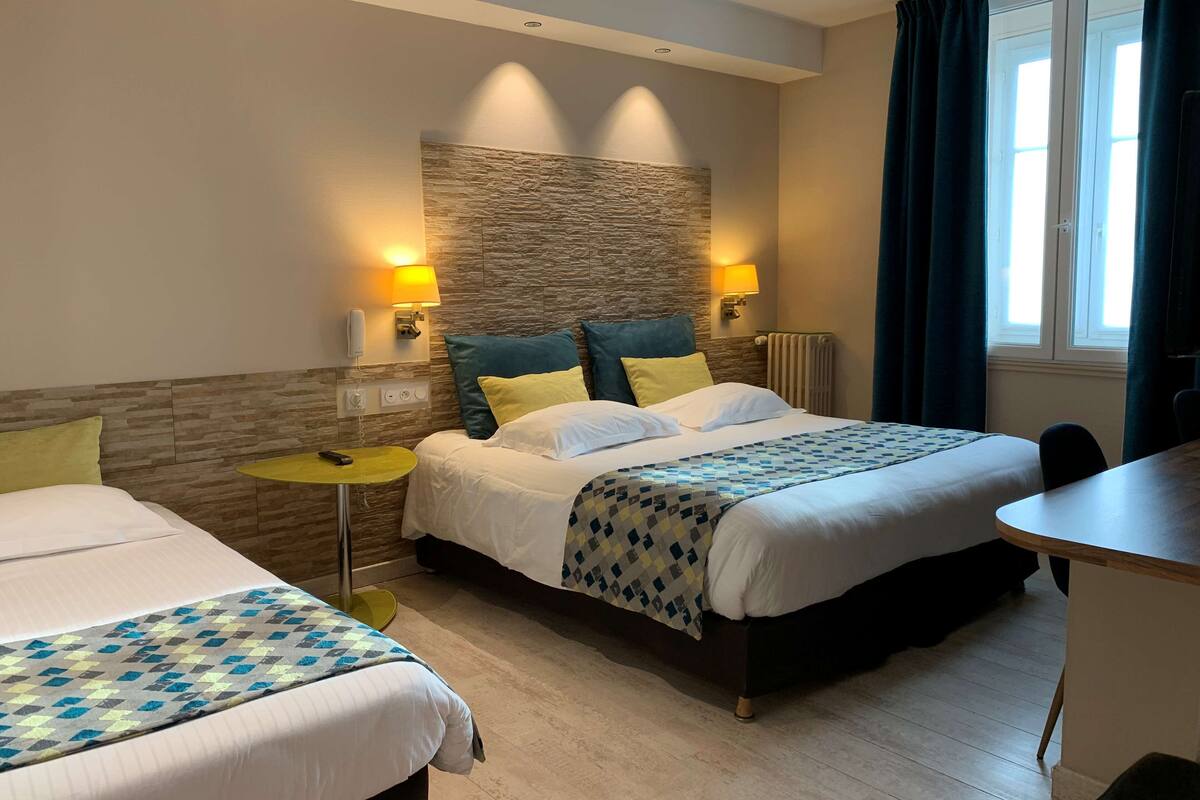 Habitación de hotel con dos camas, una mesita, una lámpara y un ventanal con cortinas.
