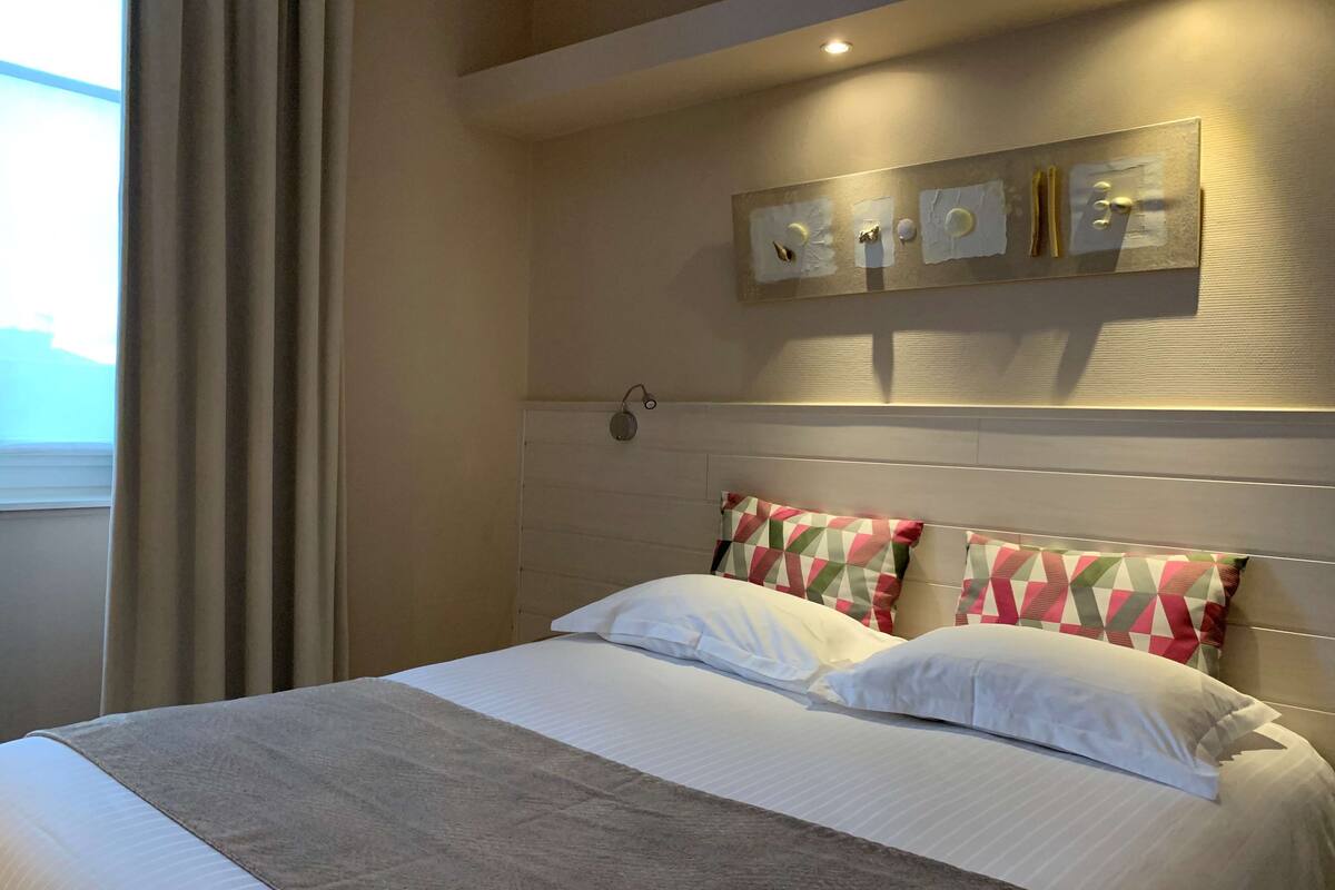 Una cama bien hecha con una manta color crema, dos almohadas estampadas y un cabecero con una lámpara de pared.