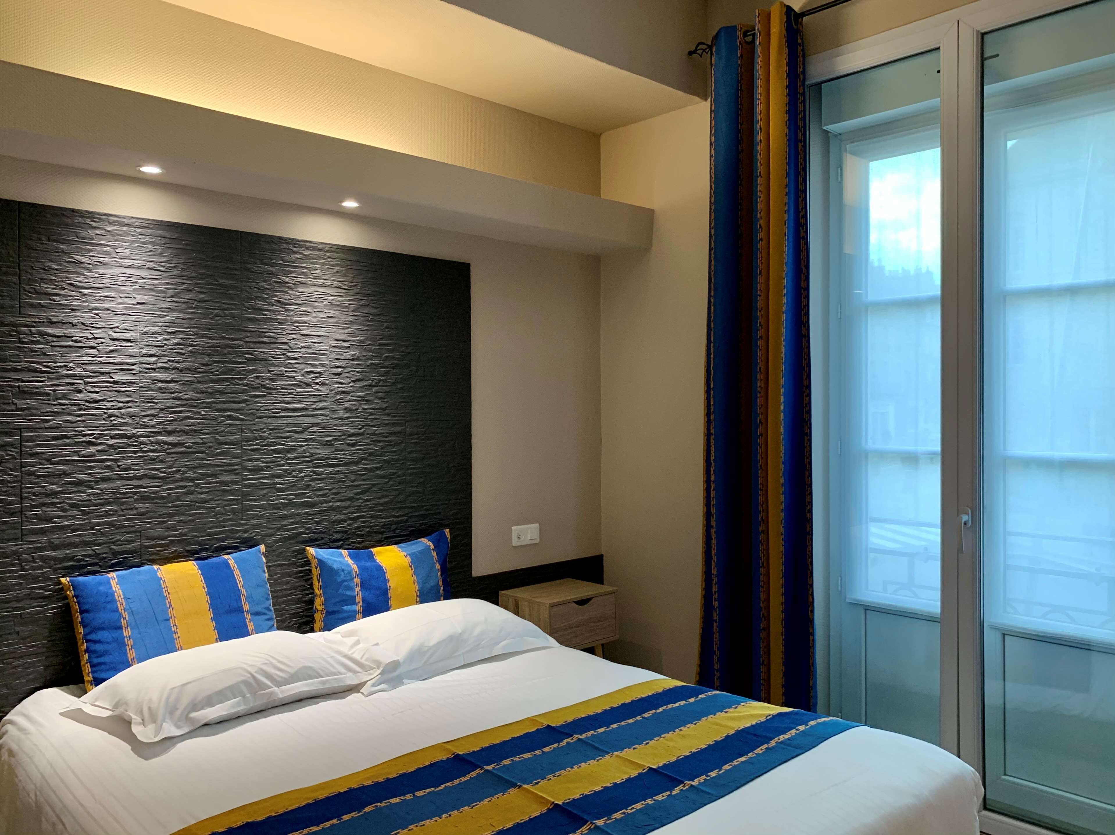 Una habitación de hotel con cama, mesita de noche y un amplio ventanal con cortinas.