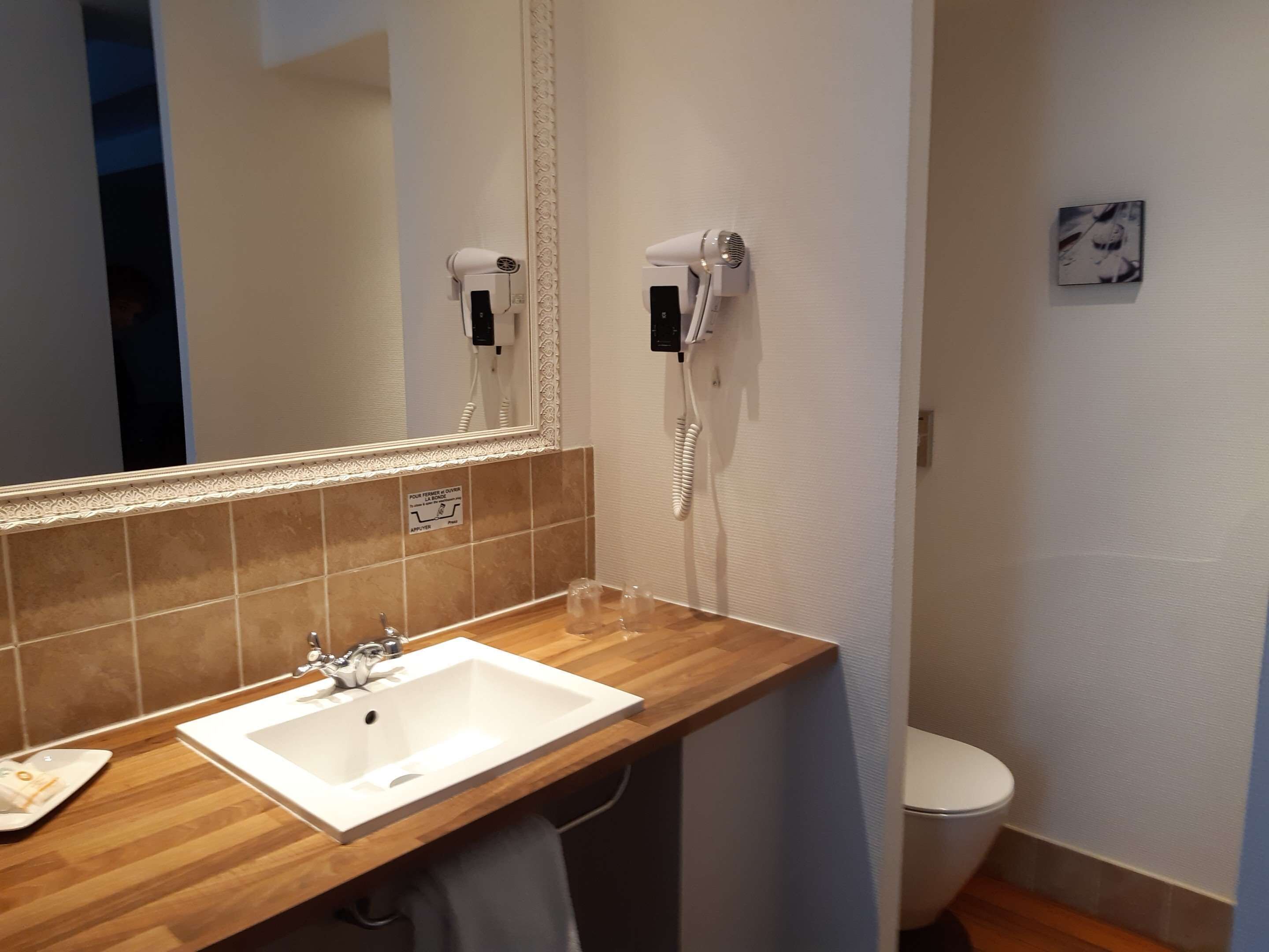 Une salle de bain avec un meuble-lavabo en bois, un lavabo blanc, un miroir avec un cadre décoratif, un sèche-cheveux et des toilettes.