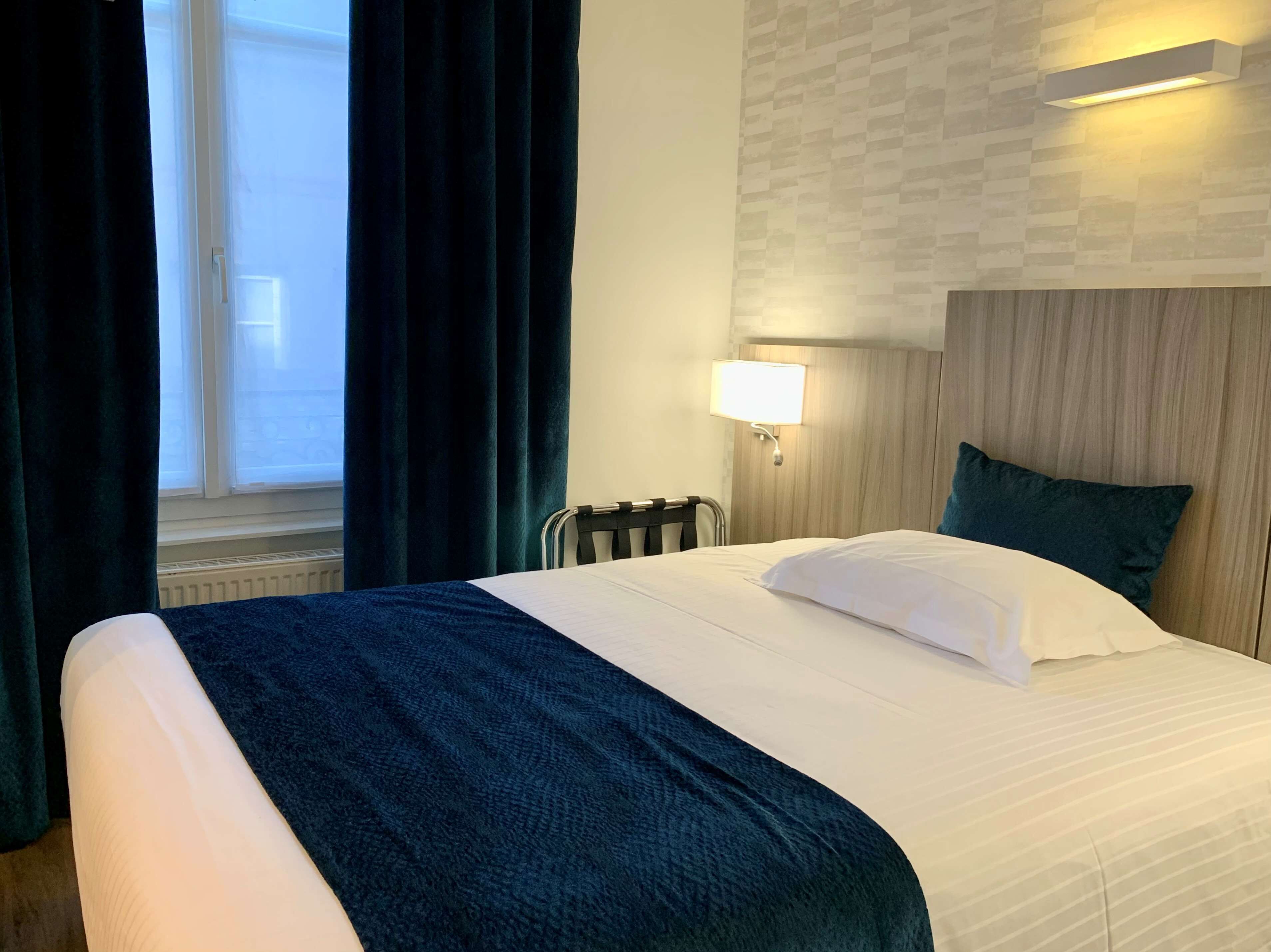 Una habitación de hotel con cama, ventana con cortinas, lámpara de pared y radiador.