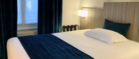 A hotel room with a bed, a window with curtains, a wall-mounted lamp, and a radiator.