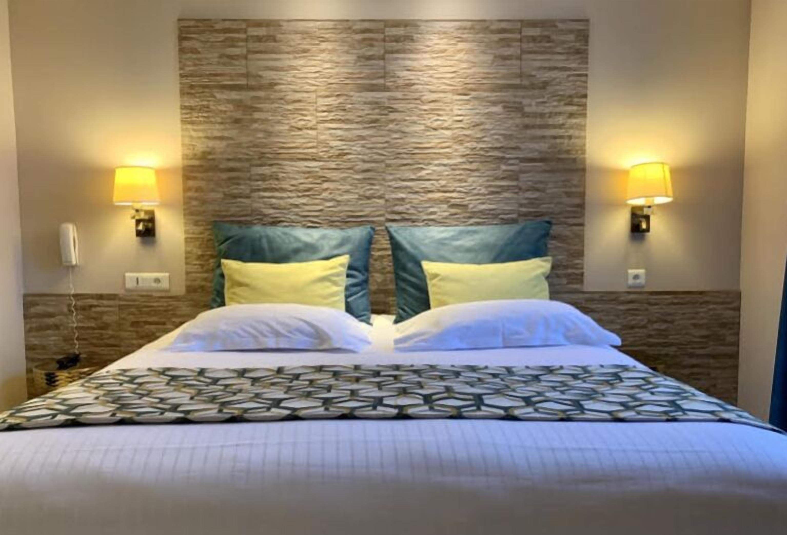 A hotel room with a bed, two wall-mounted lamps, and a textured stone wall behind the bed.