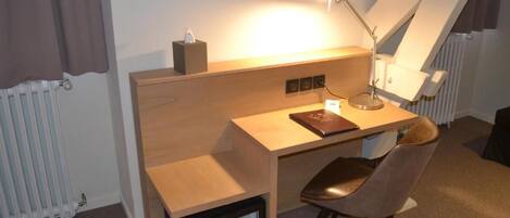 A compact bedroom with a single bed, a desk with a chair, a radiator, and a small storage unit.