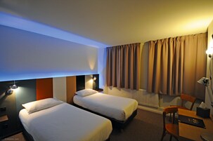 Habitación de hotel con dos camas individuales, un escritorio con silla, una lámpara y un radiador.