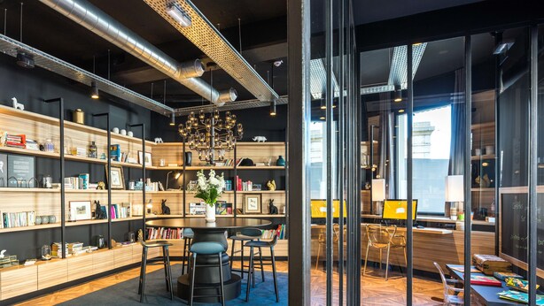 A modern library with bookshelves, a central table with chairs, and a large window providing natural light. - ibis Styles Laval Centre Gare (Laval)