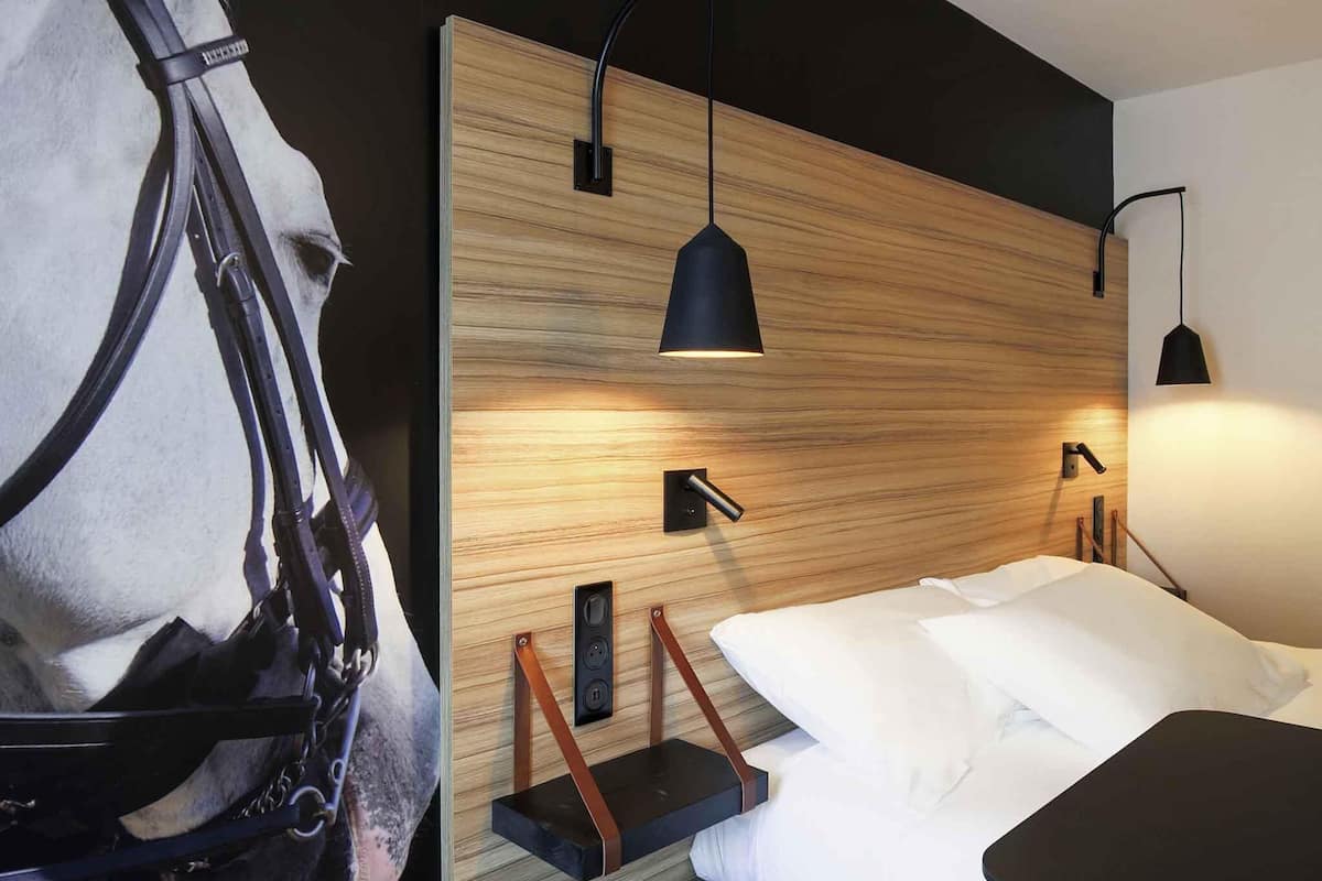 Una habitación de hotel moderna con cabecera de madera, lámparas colgantes negras, cama con ropa blanca y decoración de cabeza de caballo en la pared.
