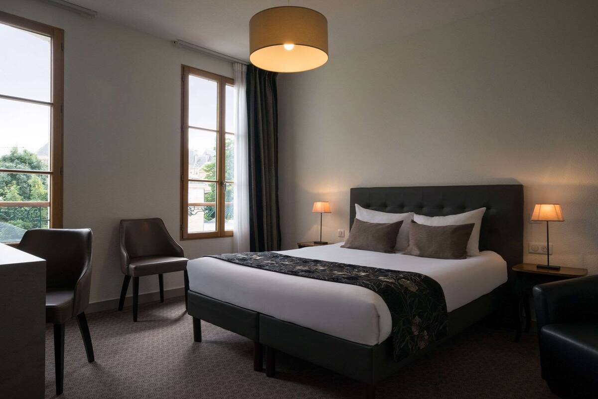 A hotel room with a bed, two chairs, a desk, a lamp, and a window with curtains.