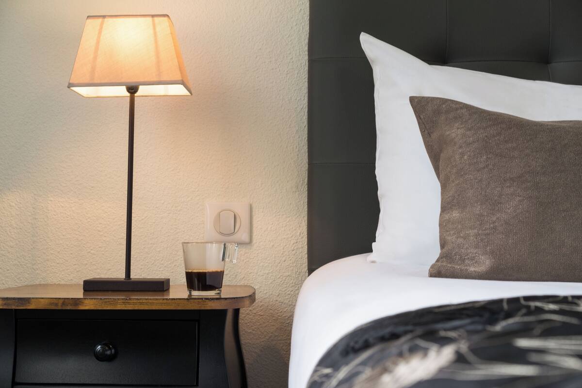 A neatly made bed with white and brown pillows, a bedside table with a lamp, and a cup of coffee.