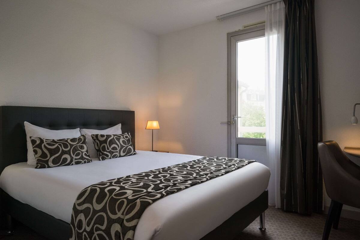 A hotel room with a bed, a desk, a chair, a lamp, and a window with curtains.