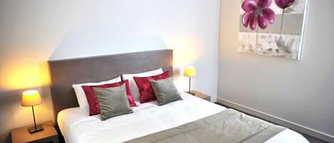 A neatly made bed with white and grey bedding, two bedside lamps, and a wall-mounted painting of flowers.