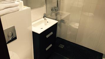 A modern bathroom with a white sink, dark floor tiles, a glass shower enclosure, and a stack of white towels.