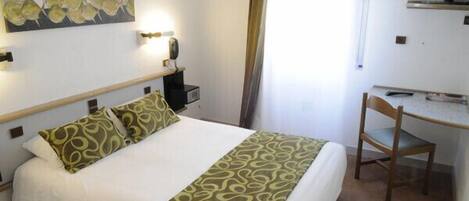 A hotel room with a bed, a desk, a chair, a painting on the wall, and a window with curtains.
