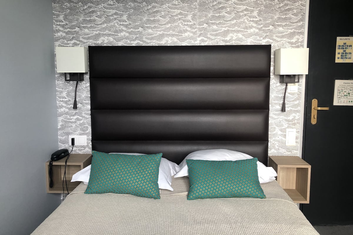 Une chambre d’hôtel avec un grand lit, deux lampes de chevet, une table de nuit en bois, un téléphone et une porte munie d’un panneau indiquant les consignes de sécurité.