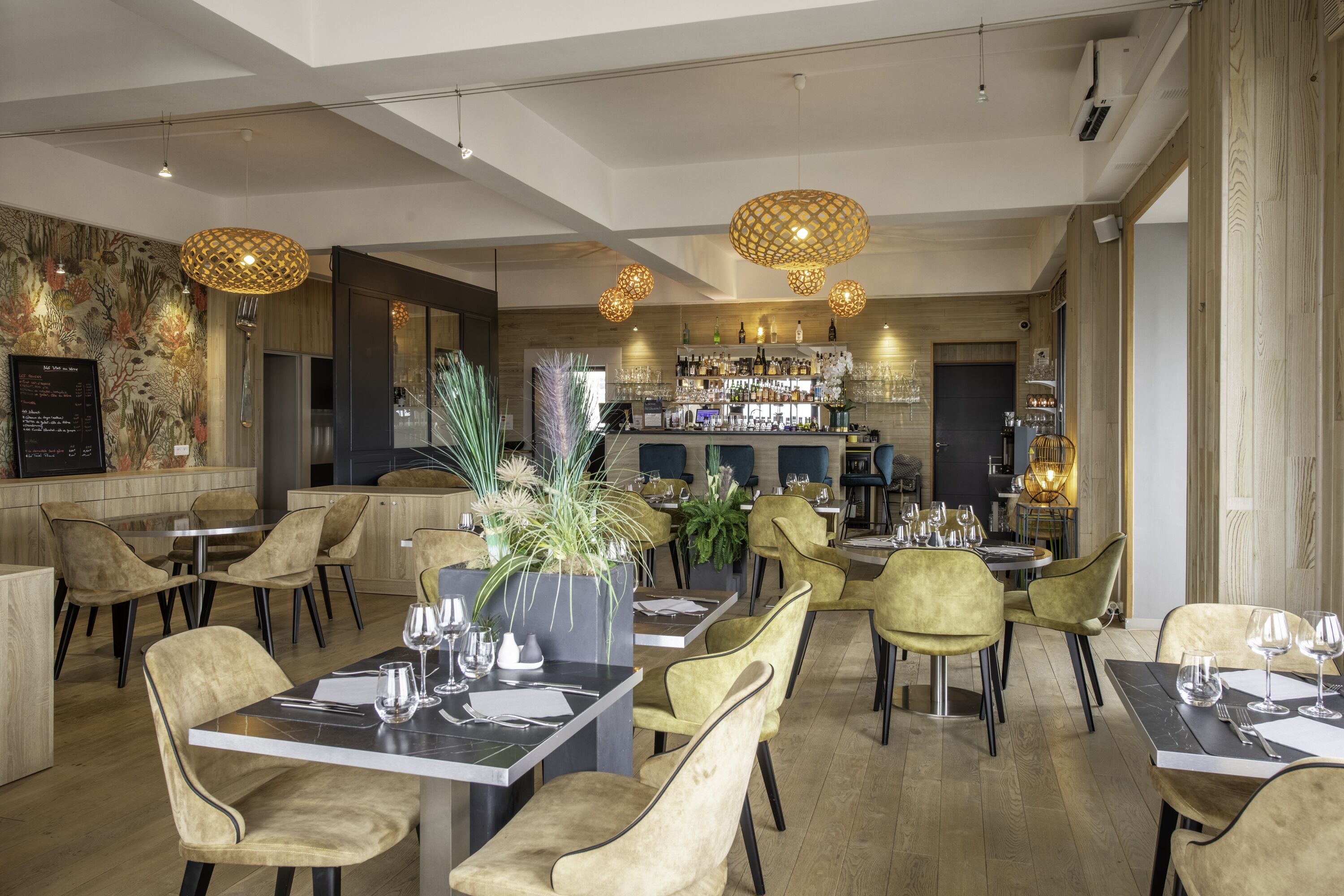 Interior de un restaurante con mesas redondas, sillas acolchadas, lámparas colgantes y una barra de fondo.