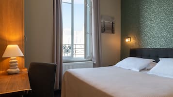 A hotel room with a bed, a desk with a lamp, a chair, a window with curtains, and a wall with a textured pattern.