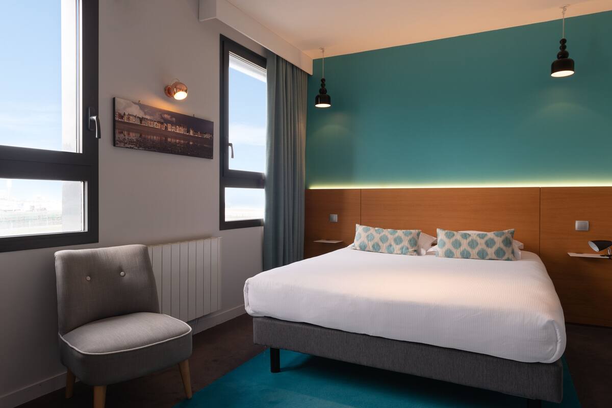 Une chambre d’hôtel moderne avec un grand lit, une chaise et une vue sur la ville par la fenêtre.
