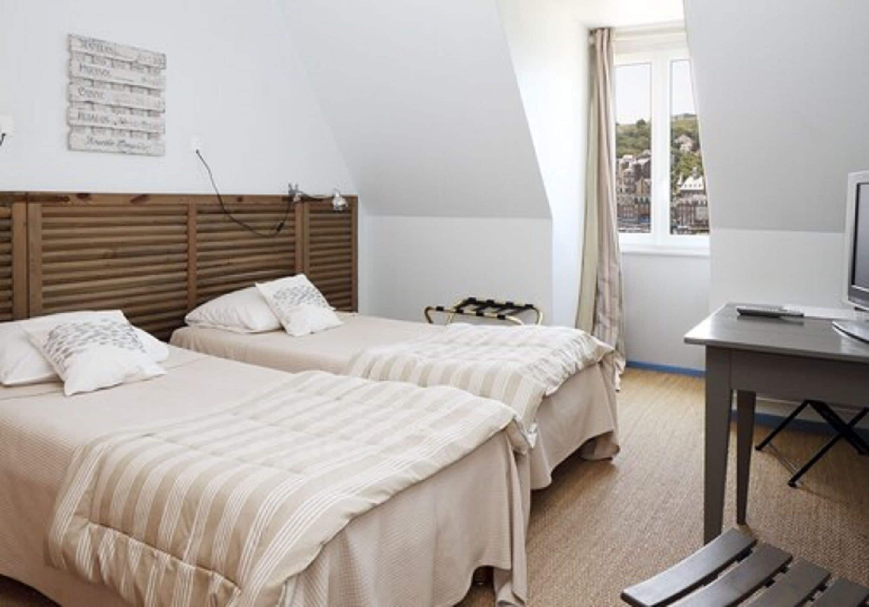 Une chambre avec deux lits, une tête de lit en bois, une petite table et une fenêtre avec des rideaux.