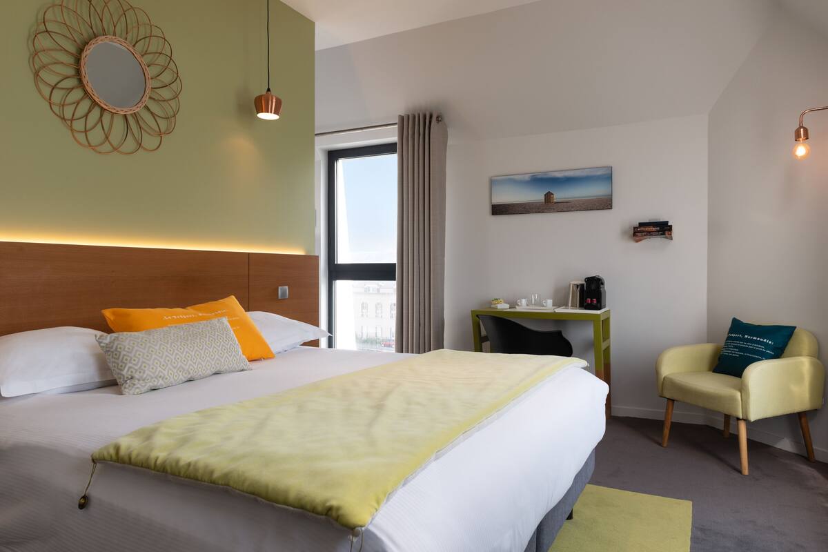 Une chambre d’hôtel avec un grand lit, un bureau, une chaise, une fenêtre avec des rideaux et un tableau au mur.