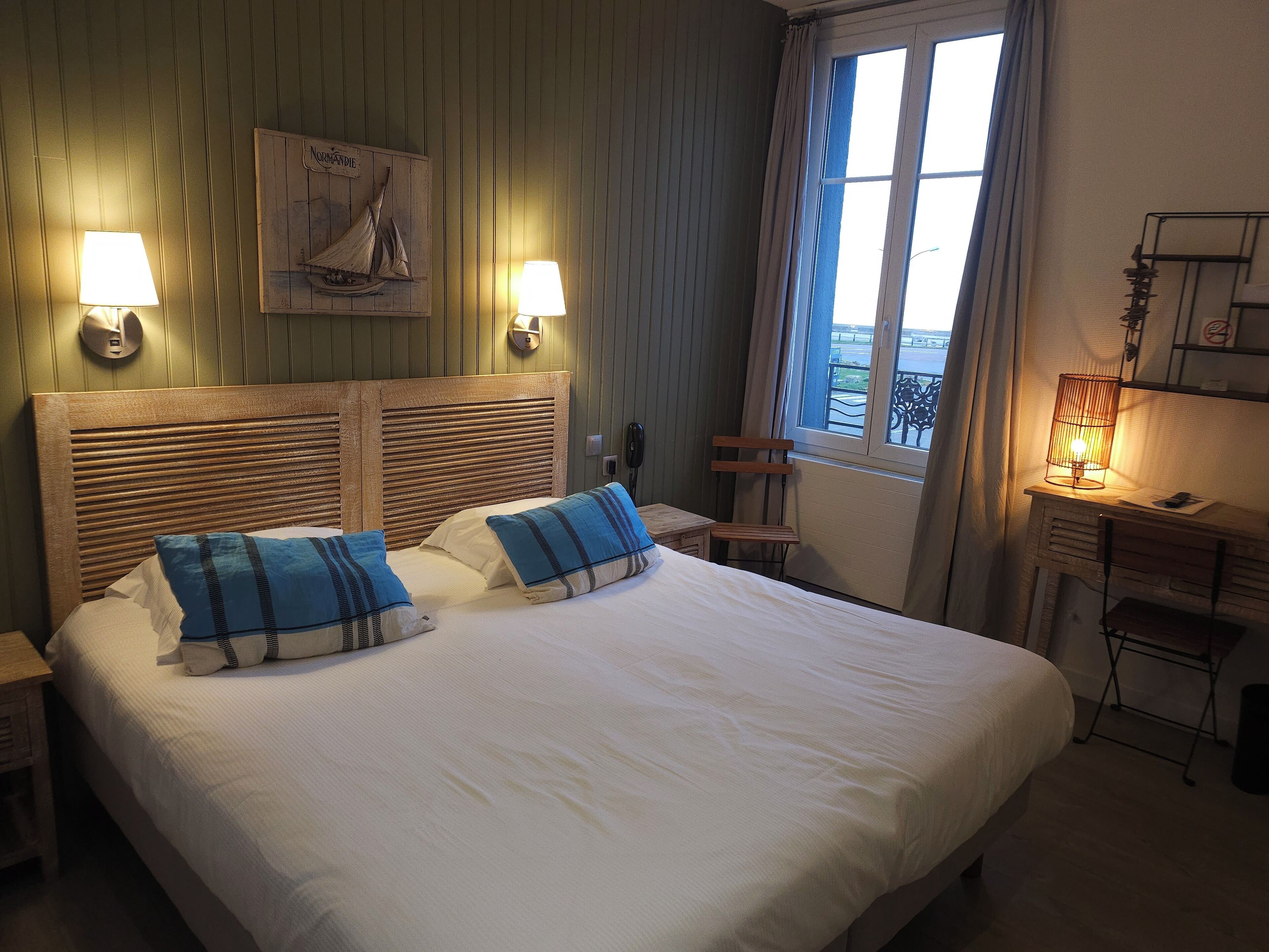 A hotel room with a large bed, two bedside lamps, a wooden headboard, a small table with a lamp, a chair, and a window with curtains.