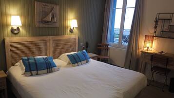 Habitación de hotel con una cama grande, dos lámparas de noche, un cabecero de madera, una mesita con lámpara, una silla y una ventana con cortinas.