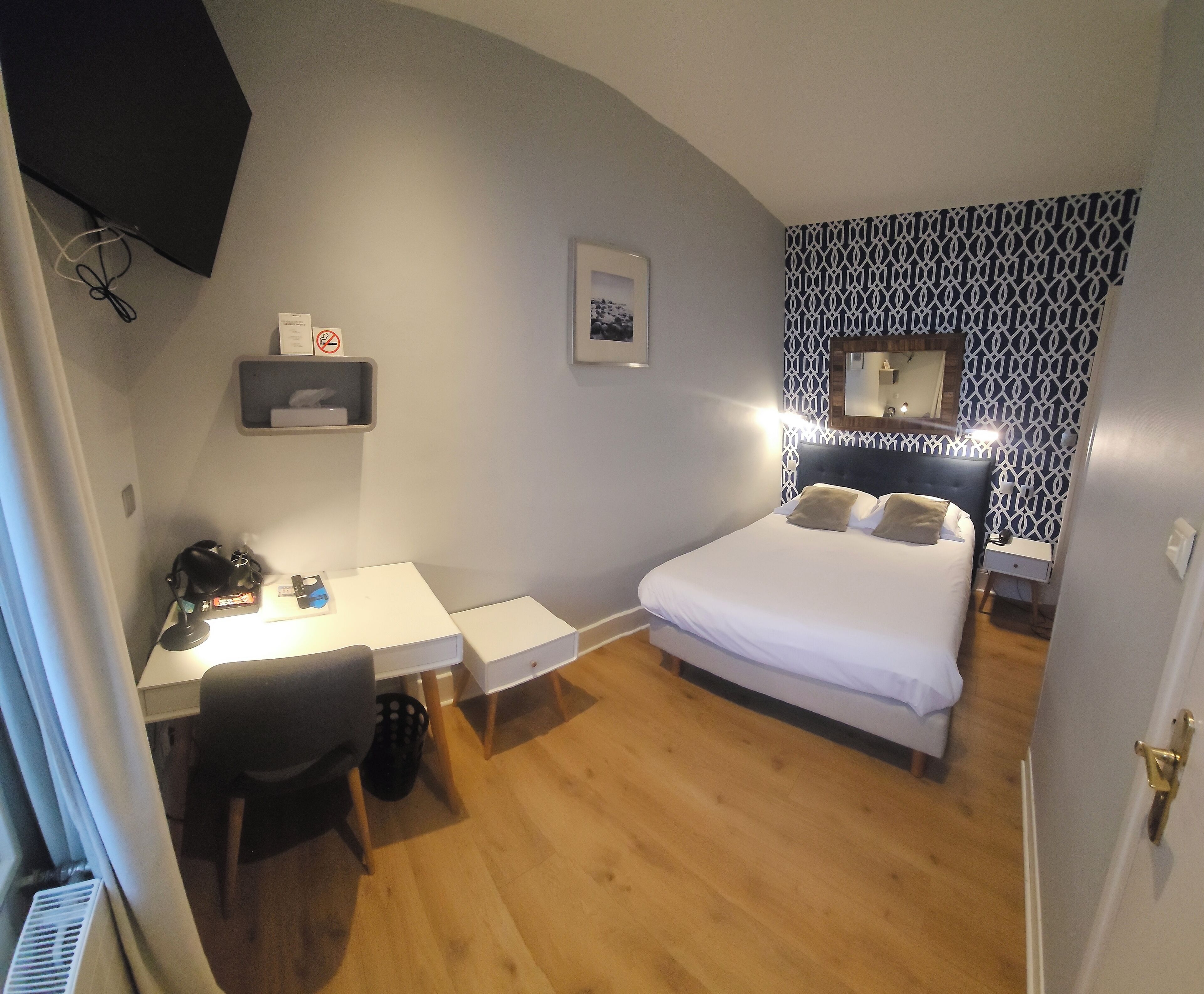 Un dormitorio con cama, escritorio y silla. Hay un estante pequeño con un cuadro enmarcado y un espejo sobre la cama.