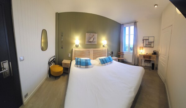 Habitación de hotel con una cama grande, un escritorio pequeño, una silla, una mesita de noche y un ventanal con cortinas.